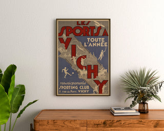 Vintage-Reiseposter – Vichy, Sport das ganze Jahr, gerahmt über einer Holzkommode zwischen Pflanzen.