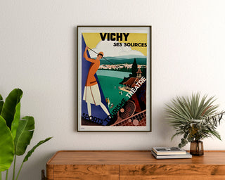 Vintage-Reiseposter – Vichy, Quellen und Tourismus, gerahmt über einer Holzkommode zwischen Pflanzen.