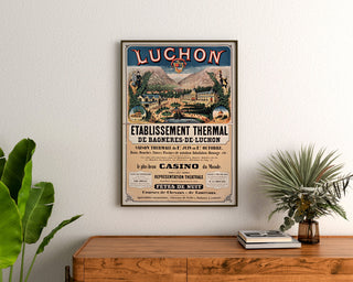 Vintage-Plakat Bagnères-de-Luchon, Kurwesen, gerahmt über einer Holzkommode, zwischen Pflanzen.